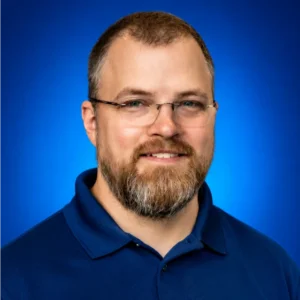 Portrait of a smiling man with glasses and a beard, wearing a blue polo against a blue backdrop.
