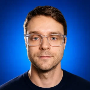 Evgeny Tkachenko Portrait of a man wearing clear-framed glasses against a blue studio background.