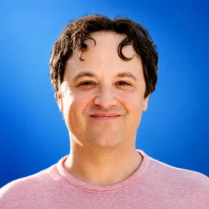Smiling man facing the camera against a solid blue background, wearing a pink shirt.