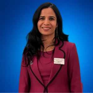 Harpreet Kaur Gill Professional portrait of a woman with long dark hair, wearing a magenta blazer and a name badge, smiling against a blue gradient backdrop.