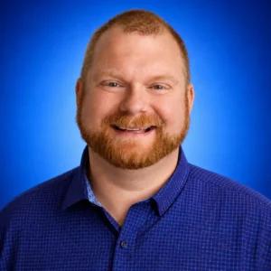 Head-and-shoulders portrait of a smiling man with a red beard wearing a blue checkered shirt against a blue studio backdrop.