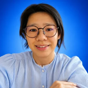 Juliet Lim Woman with short dark hair and round glasses smiling at the camera, blue background.