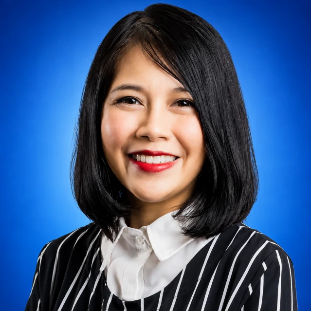 Smiling woman with shoulder-length black hair in a white collared shirt and black striped blazer, against a blue gradient background.