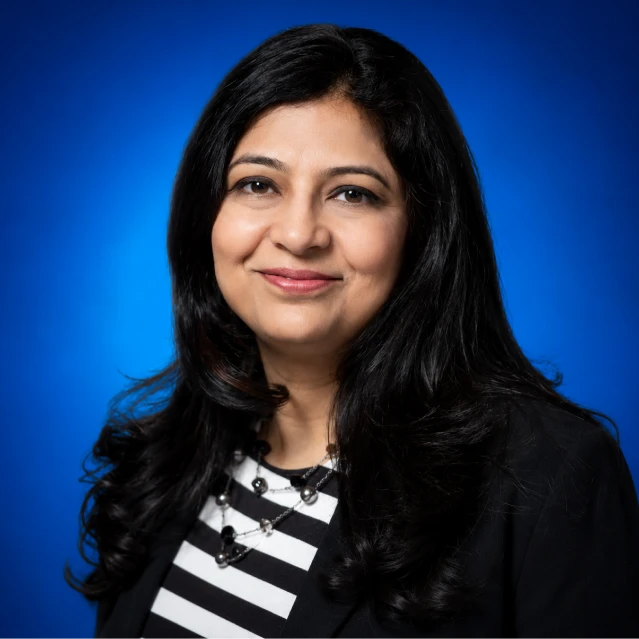 Professional headshot: smiling woman with long black hair, wearing a black blazer over a black-and-white striped top, against a blue gradient background