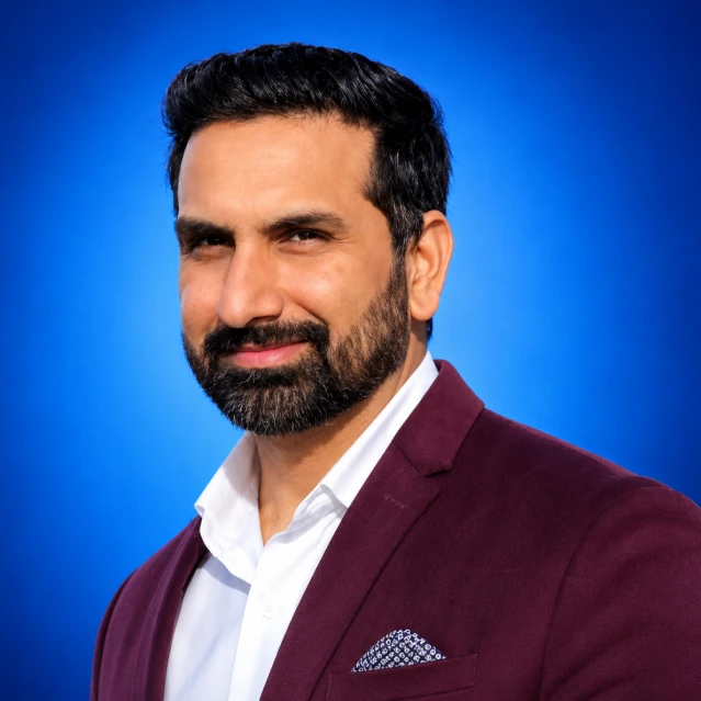 Professional headshot of a bearded man in a burgundy blazer and white shirt, with a blue gradient backdrop and pocket square in the breast pocket.