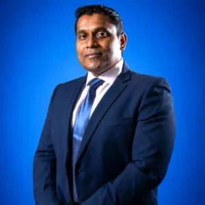Professional portrait of a man in a navy suit and blue tie, standing against a blue studio backdrop, looking confident.