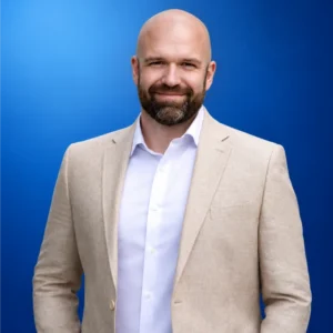 Steve Leggett Smiling bald man with a beard in a beige blazer and white shirt, posing against a blue gradient background.