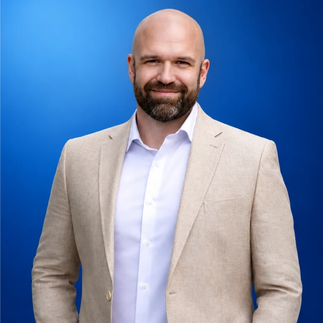 Smiling bald man with a beard in a beige blazer and white shirt, posing against a blue gradient background.