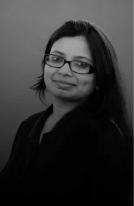 Portrait of a smiling woman with glasses and long dark hair, wearing a black top against a plain gray background.