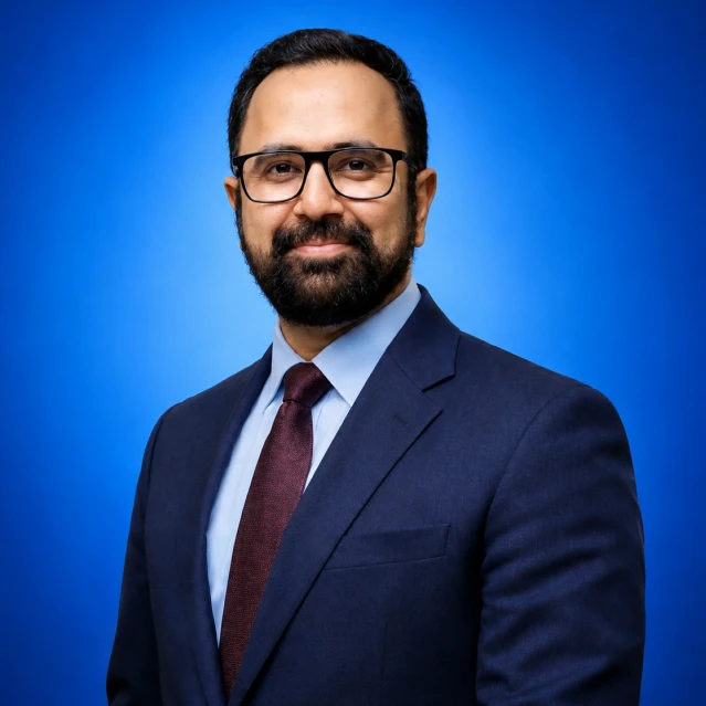 Professional headshot of a man with glasses and a beard, wearing a navy suit, light blue shirt, and burgundy tie against a blue backdrop.
