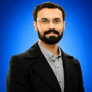 Portrait of a man with glasses and a beard, wearing a gray button-down shirt and black blazer, against a blue backdrop.