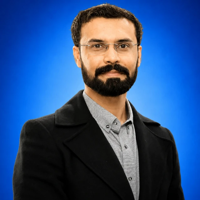 Portrait of a man with glasses and a beard, wearing a gray button-down shirt and black blazer, against a blue backdrop.
