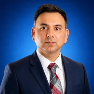 Professional head-and-shoulders portrait of a man in a navy suit, white shirt, and red plaid tie against a blue background, looking at the camera.