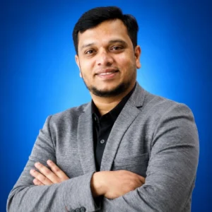 Ashwin Balaji Kuppuraj Portrait of a man in a gray blazer with a black shirt, arms crossed, smiling against a blue backdrop.