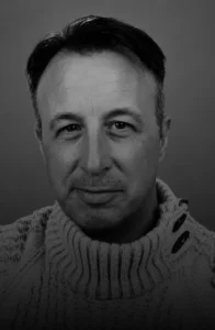 Black-and-white portrait of a man with short dark hair wearing a chunky knit sweater, facing the camera against a plain backdrop.