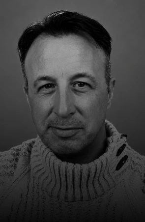 Black-and-white portrait of a man with short dark hair wearing a chunky knit sweater, facing the camera against a plain backdrop.