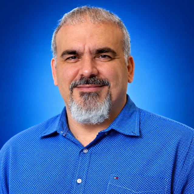 Portrait of a middle-aged man with a gray beard in a blue patterned shirt against a blue gradient backdrop.