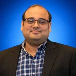 Feroz Louis Portrait of a man in a black blazer and blue checkered shirt wearing glasses, against a blue gradient background.