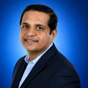 Gaurav Shah (1) Portrait of a man in a navy blazer and light blue shirt, smiling, against a blue gradient background.