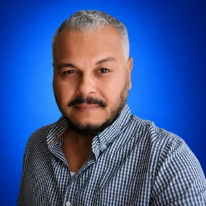 Hector Claudio Portrait of a man with short gray hair and beard, wearing a checkered shirt, against a blue backdrop.