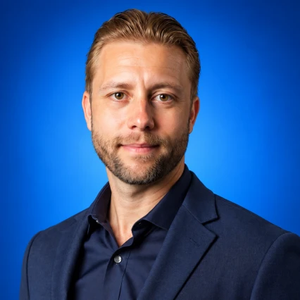 Portrait of a man with short blond hair and light beard, wearing a navy blazer against a solid blue background to convey a professional headshot.