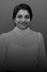 Jyothi Murala Portrait of a smiling woman with dark hair, wearing a sheer white blouse with a high ruffled collar.