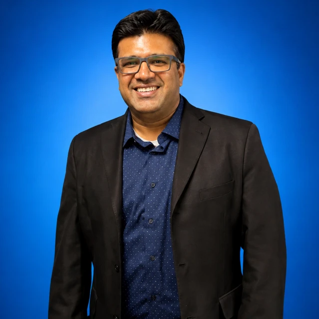 Smiling man in a dark blazer and blue patterned shirt against a blue gradient background.