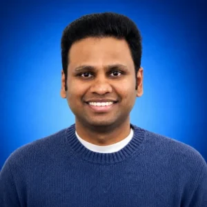 Portrait of a man with short black hair smiling, wearing a blue knit sweater against a blue gradient background.