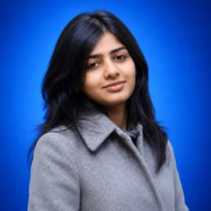 Portrait of a woman with shoulder-length dark hair wearing a light gray coat against a blue studio backdrop, looking at the camera with a slight smile.