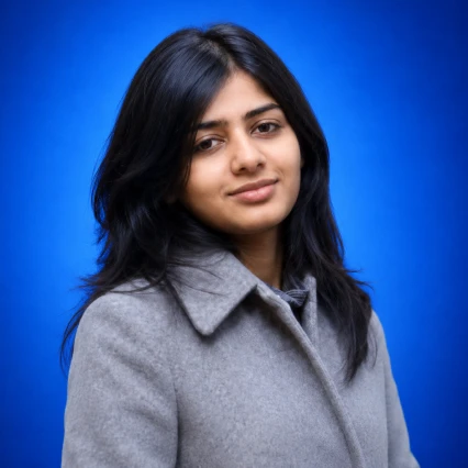 Portrait of a woman with shoulder-length dark hair wearing a light gray coat against a blue studio backdrop, looking at the camera with a slight smile.