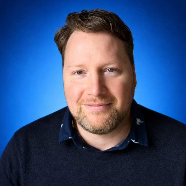 Professional headshot of a smiling man with short brown hair and light beard, wearing a navy sweater over a collared shirt, against a blue gradient backdrop.