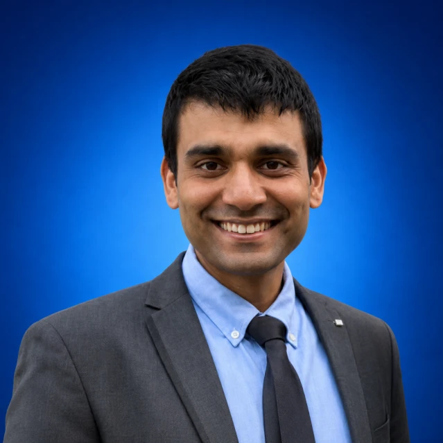 Smiling man in a gray suit and blue shirt against a blue gradient background, portrait headshot.