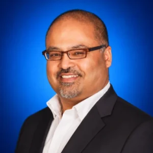 Professional headshot of a man with glasses in a black suit and white shirt, blue gradient background.