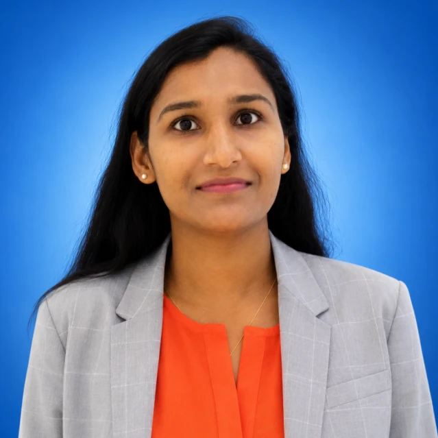 Portrait of a woman with long dark hair wearing a light gray blazer and orange blouse, against a blue backdrop.