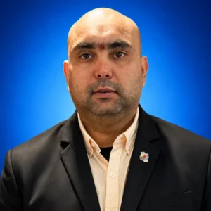 Portrait of a bald man in a beige shirt and black blazer against a blue background, wearing a small lapel pin on the left lapel.
