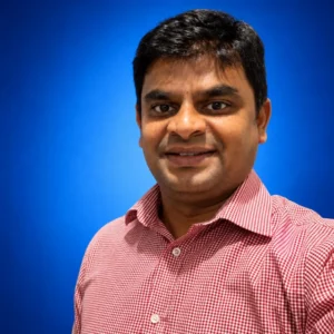 Portrait of a man with short black hair wearing a red checkered shirt, smiling against a blue backdrop.
